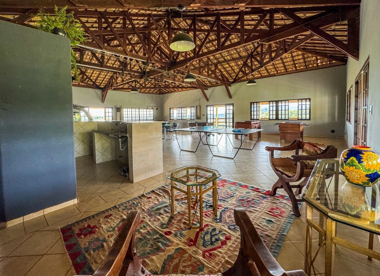 Quarto espaçoso com teto de madeira aparente. A mobília inclui tapetes, mesas, cadeiras e janelas.