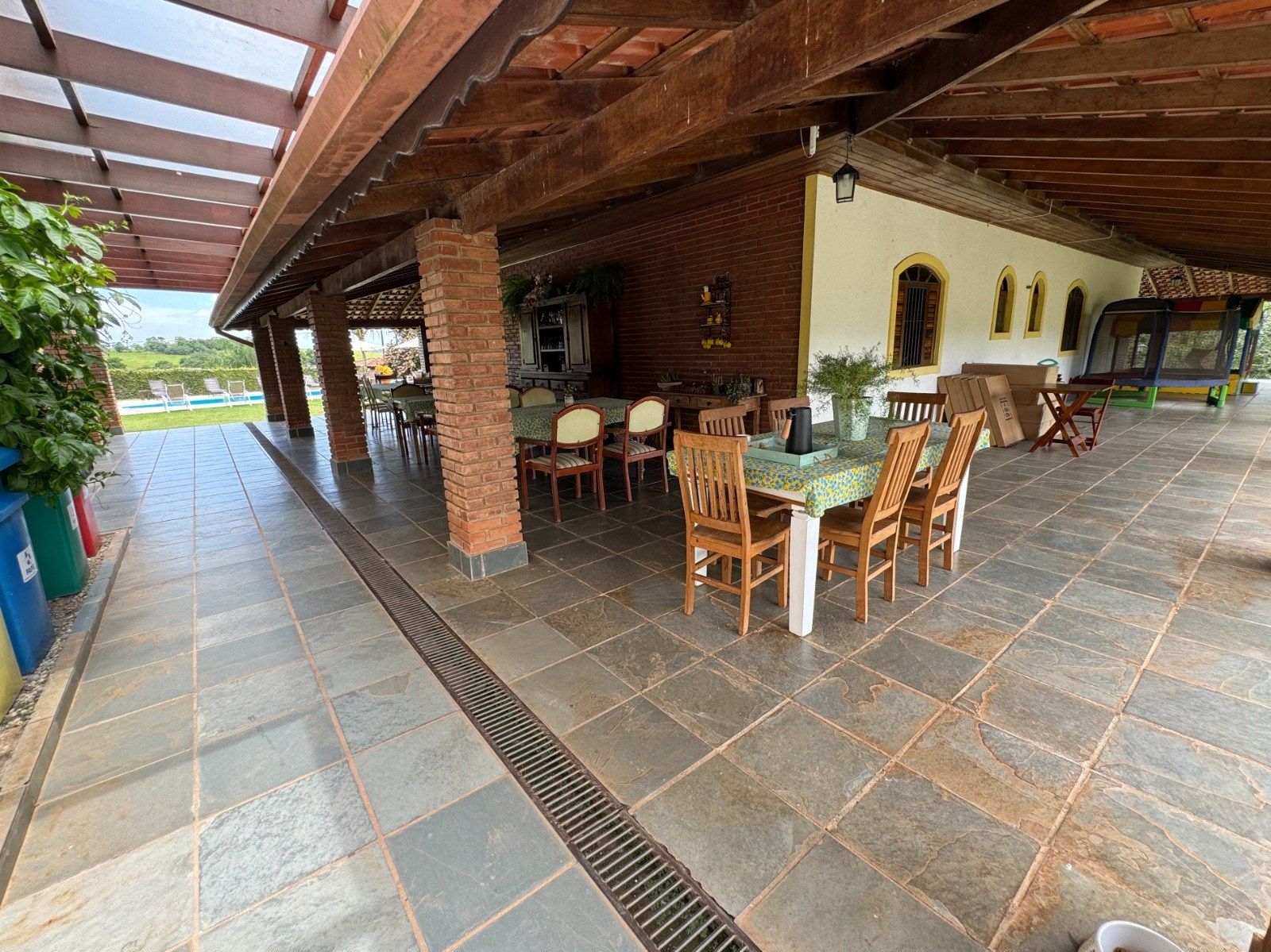 Pátio externo coberto com mesa e cadeiras para refeições, colunas de tijolos e piso de pedra.