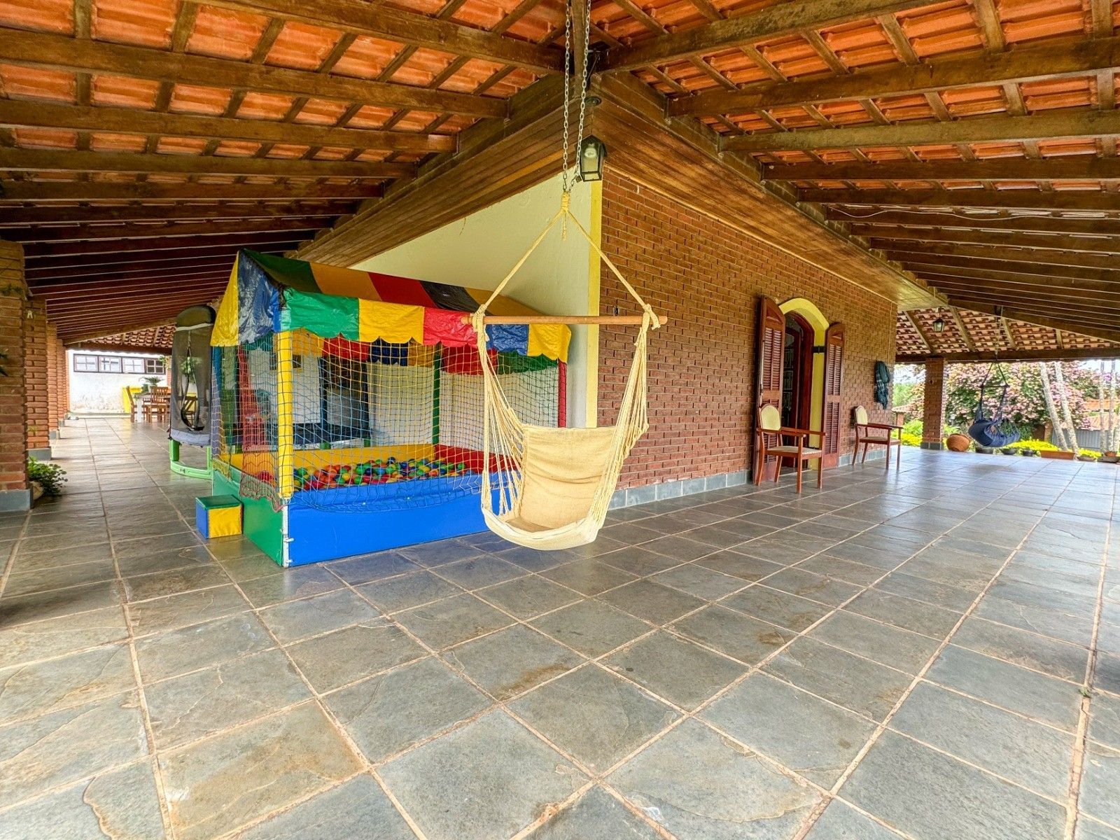 Pátio coberto com paredes de tijolos, piso de azulejo e uma cadeira suspensa perto de um brinquedo de parque infantil.