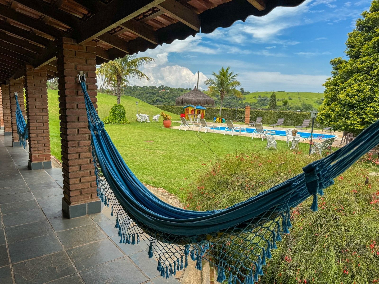 Rede azul pendurada sob um alpendre, com vista para uma piscina e campos verdes num dia ensolarado.