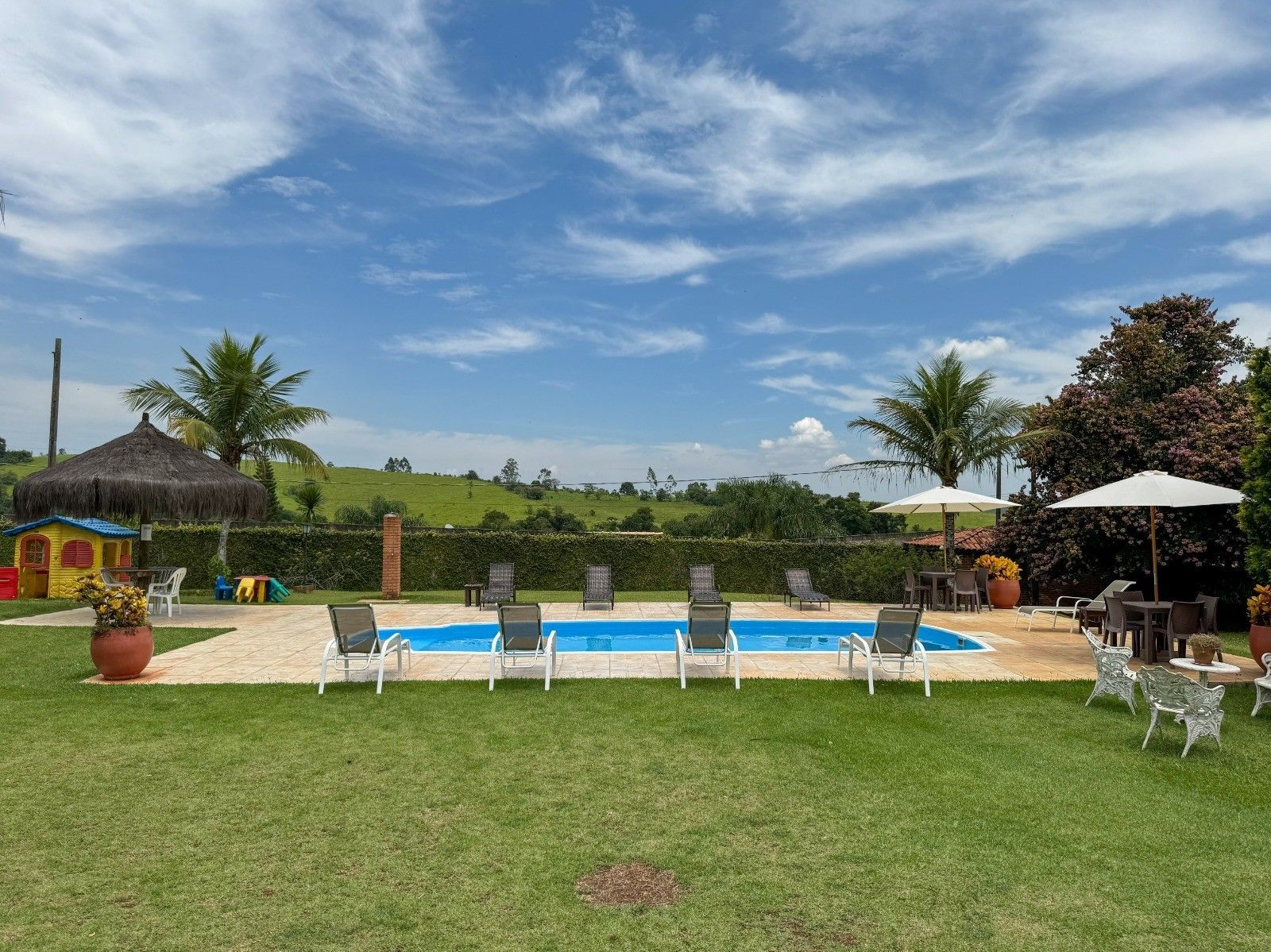 Área da piscina com cadeiras, uma pequena casinha de brincar e um gramado ensolarado sob um céu azul.