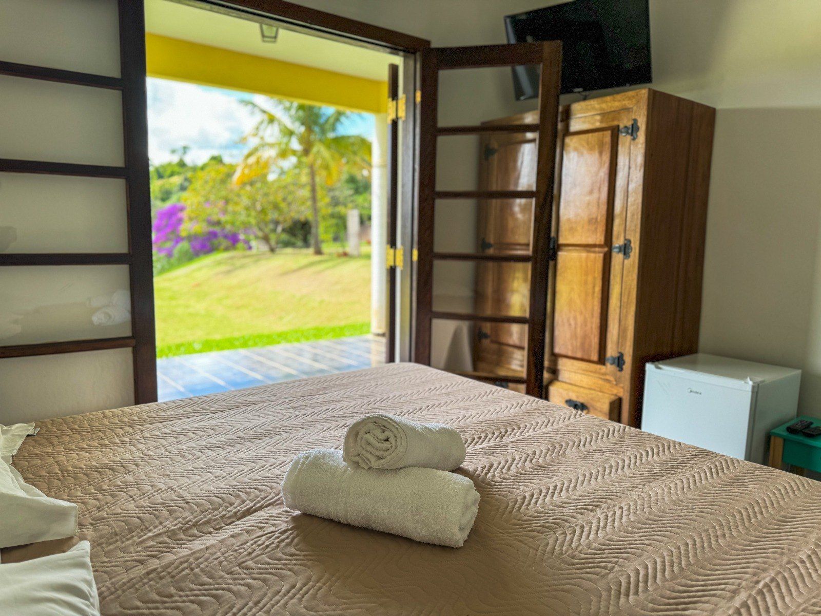 Quarto com cama, toalhas e portas abertas para um jardim. Inclui guarda-roupa de madeira e frigobar.