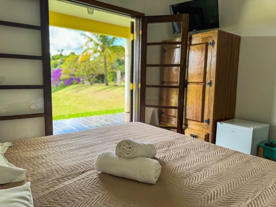 Quarto com cama, toalhas e portas abertas para um jardim. Inclui guarda-roupa de madeira e frigobar.