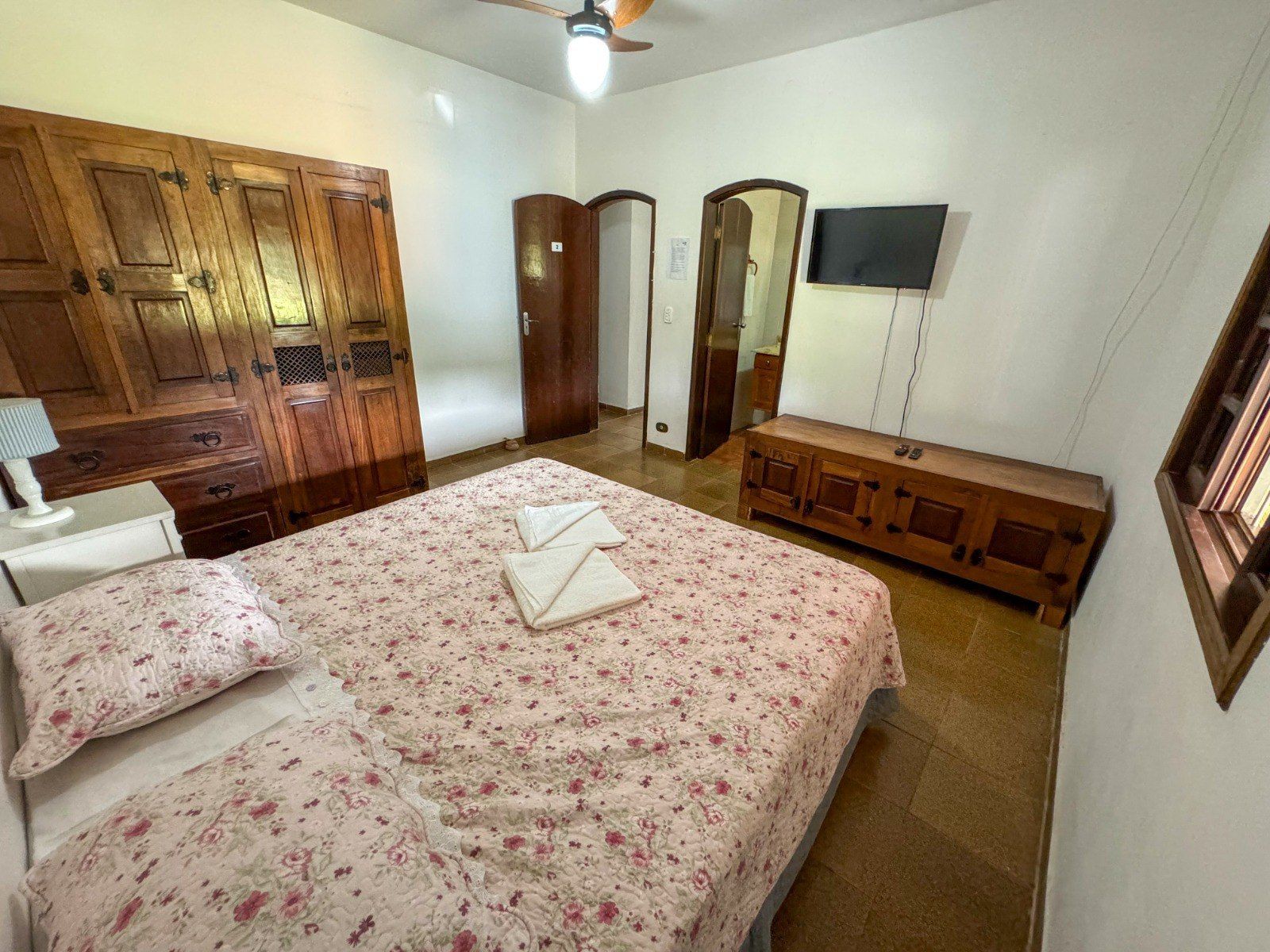 Quarto com móveis de madeira, cama com roupa de cama floral, pequena TV sobre a cômoda, porta para o banheiro.