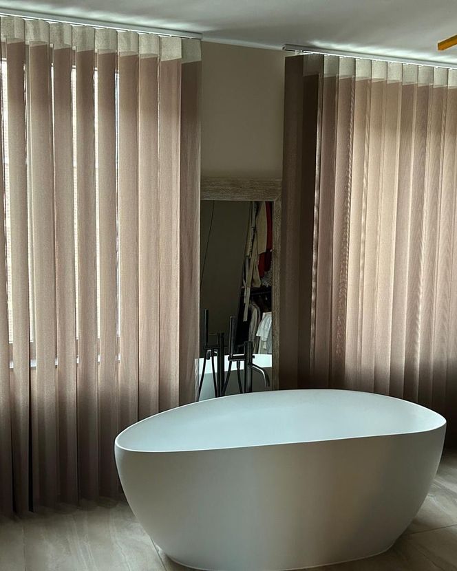 Bagno moderno con vasca ovale bianca tra tende beige e specchio a parete.