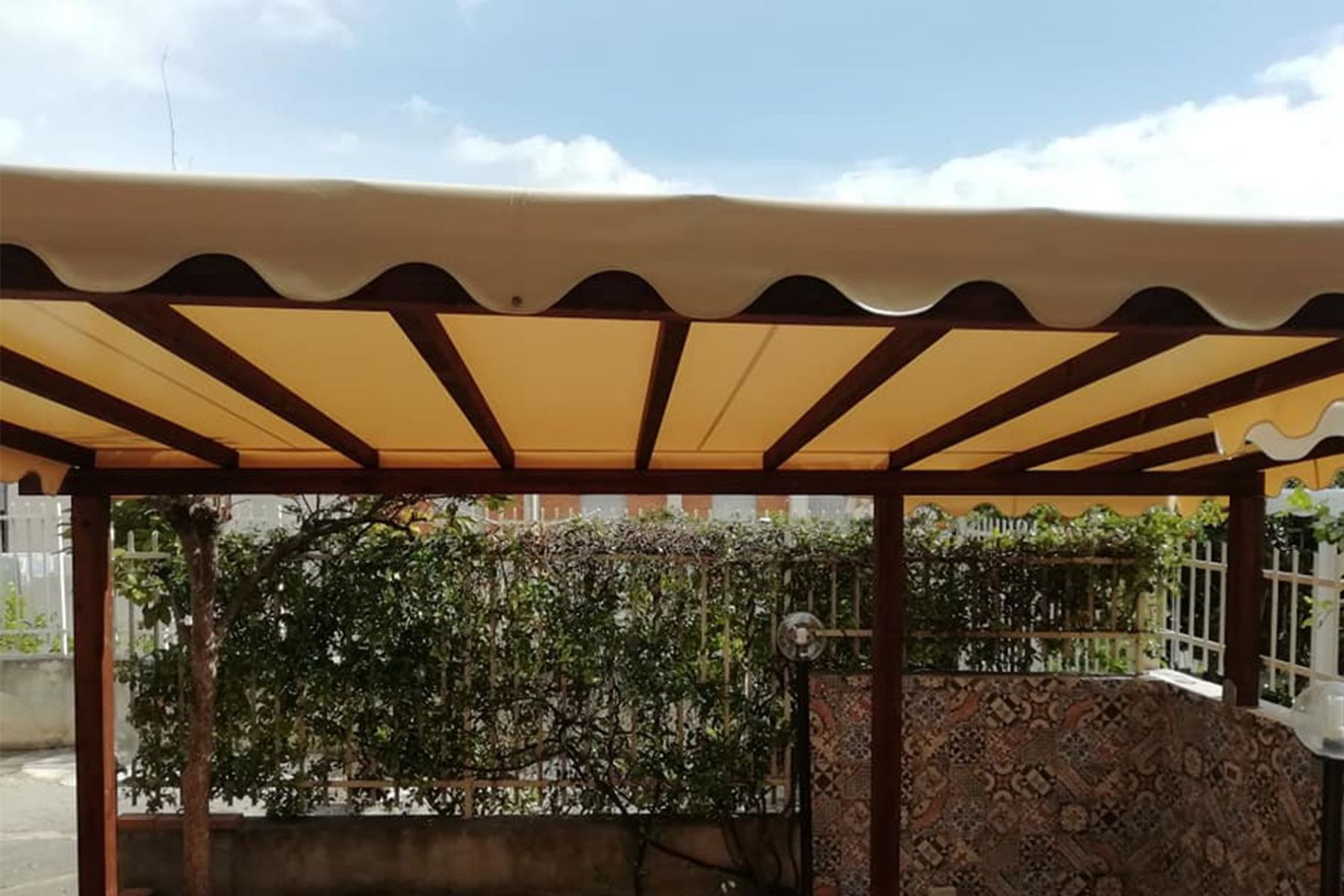 Tenda da sole a righe beige sopra una zona salotto esterna ombreggiata con piante e pannelli a parete decorati.