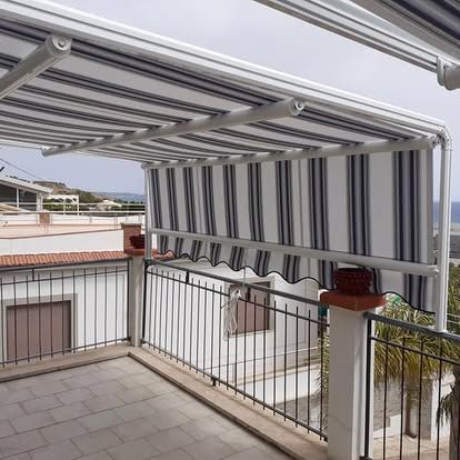 Balcone con tenda da sole retrattile a righe, ringhiera e pavimento piastrellato con vista sugli edifici circostanti.