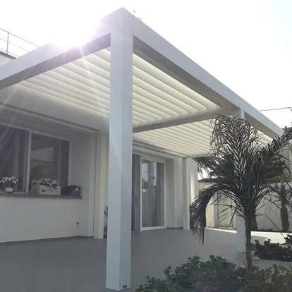Veranda moderna di una casa bianca con tettoia a pergola e palma in vaso, immersa nella luce del sole.