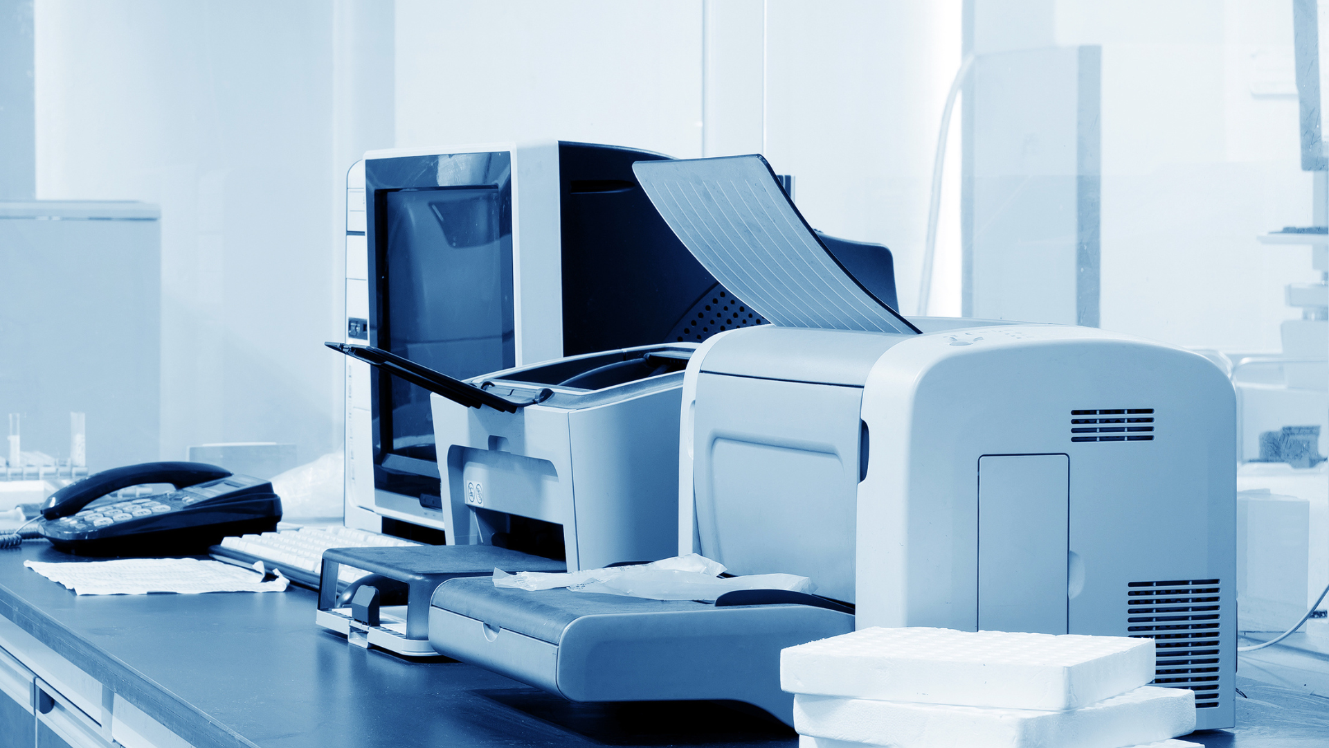 Office printers on a desk, with paper outputting. Blue-toned office setting.