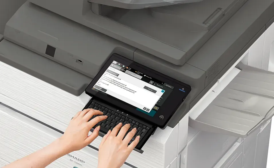 Hands typing on a keyboard attached to a multifunction printer, with a touch screen displaying text.