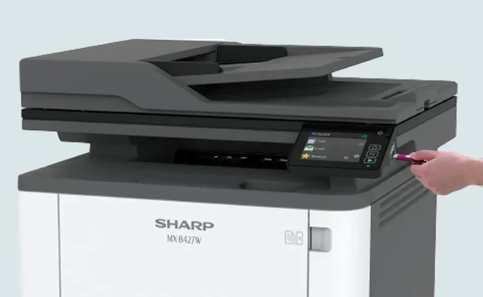 Hand inserting a USB drive into a Sharp multifunction printer. The printer is white and gray.