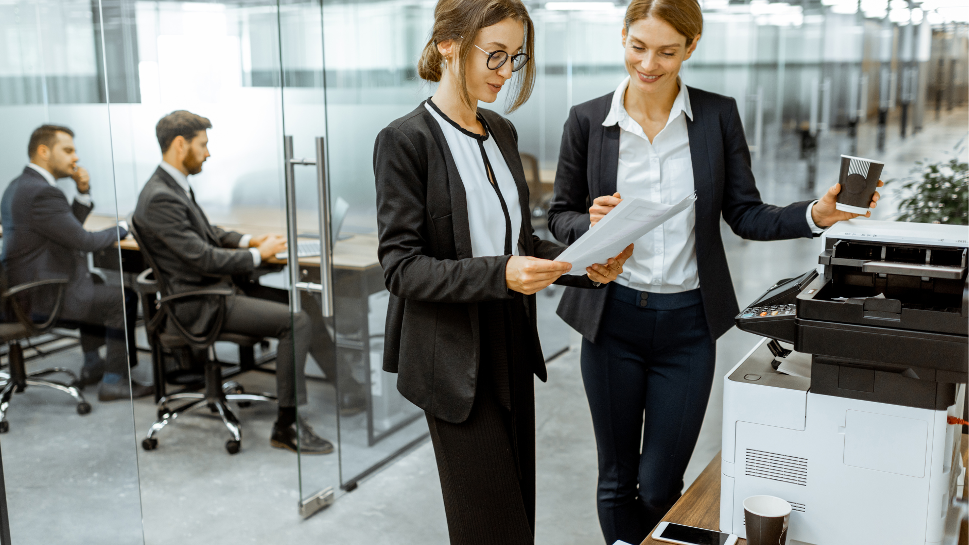 Two women in suits reviewing documents near a printer in an office.