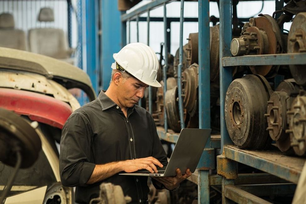 A man in a hard hat uses a laptop in a salvage yard, surrounded by car parts and equipment.