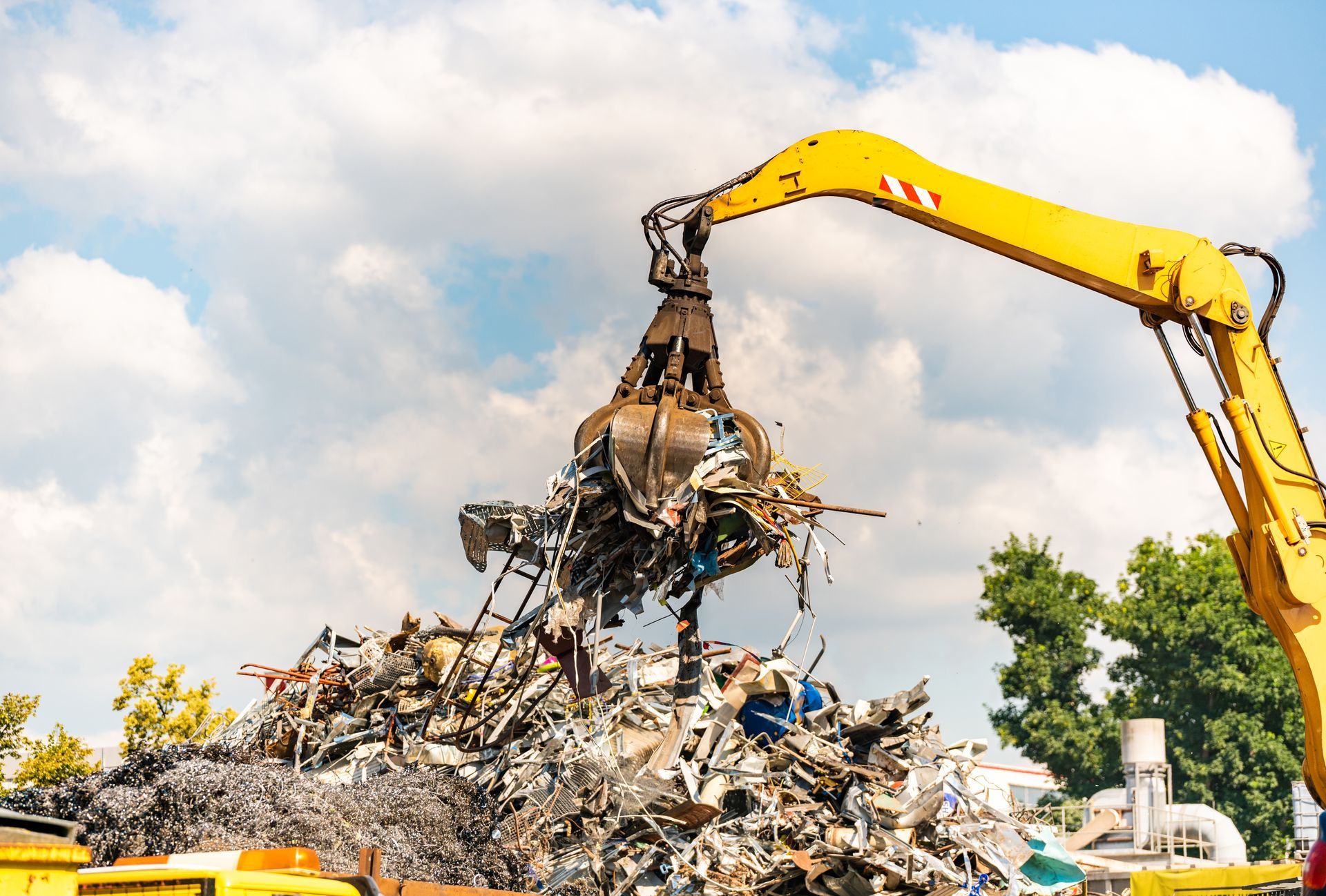 A yellow excavator with a claw lifts scrap metal from a large pile against a cloudy sky.