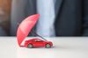 Red toy car protected by a small red umbrella, symbolizing insurance. A person in a suit is in the background.
