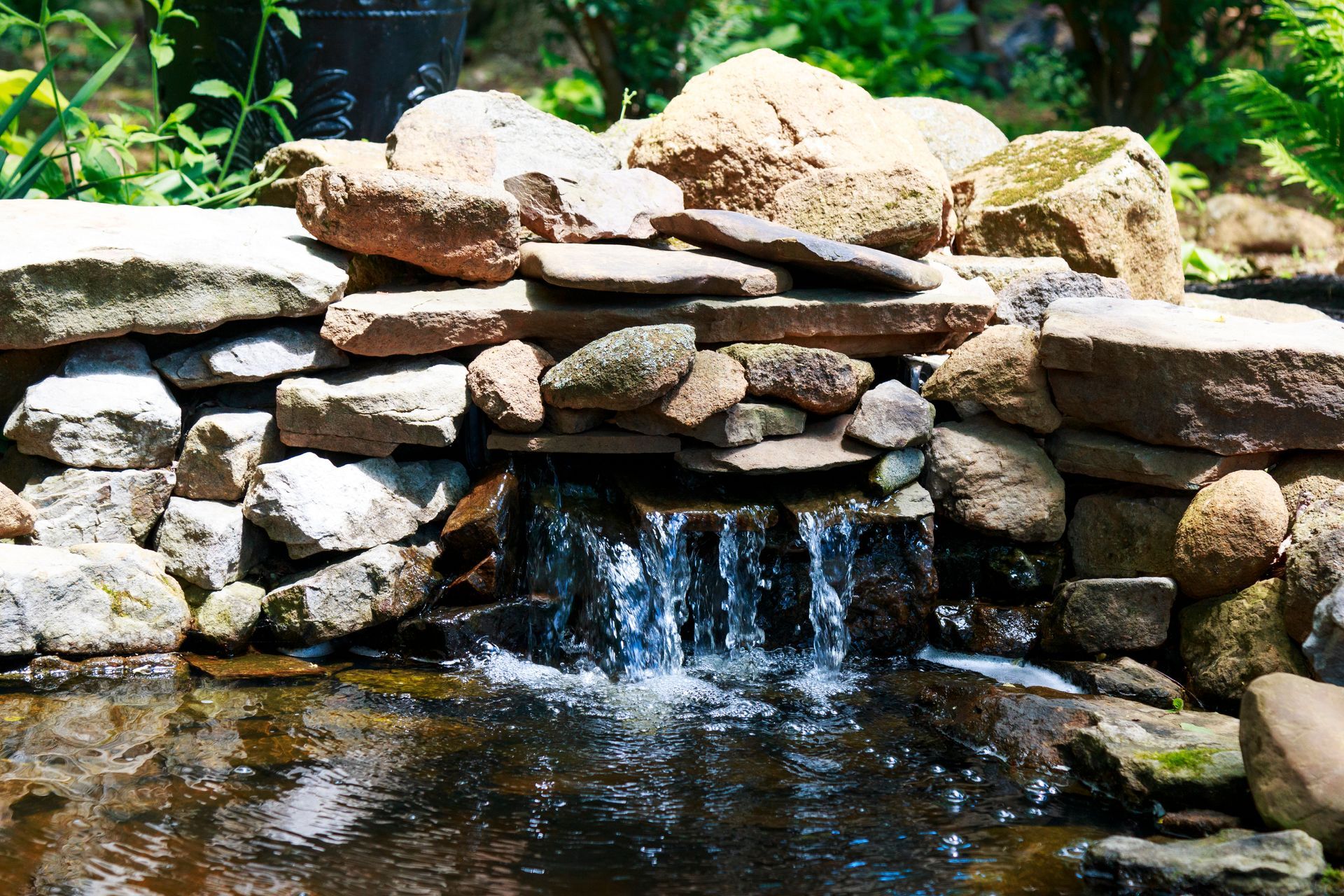 Small stone waterfall flowing through a stacked rock garden with greenery behind it