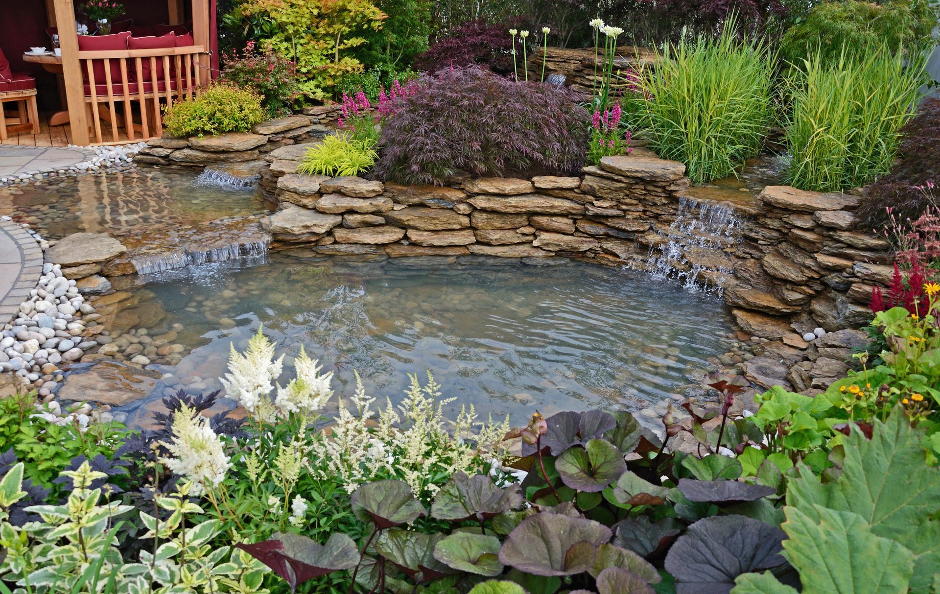 Small garden pond with stone edging, surrounded by lush plants and flowers