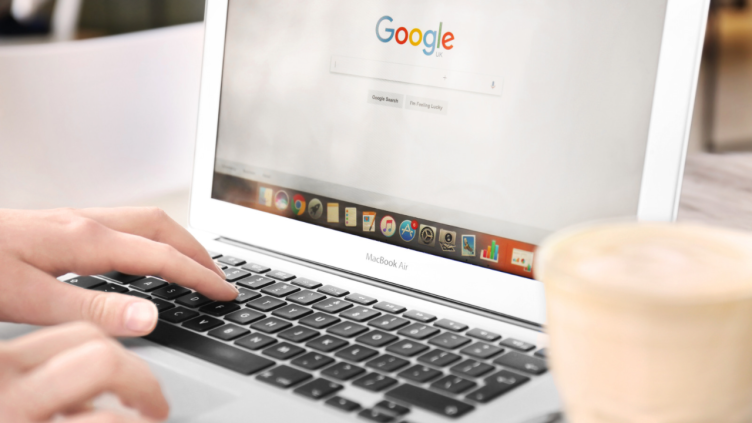 Uma pessoa está digitando em um laptop com uma xícara de café ao fundo.