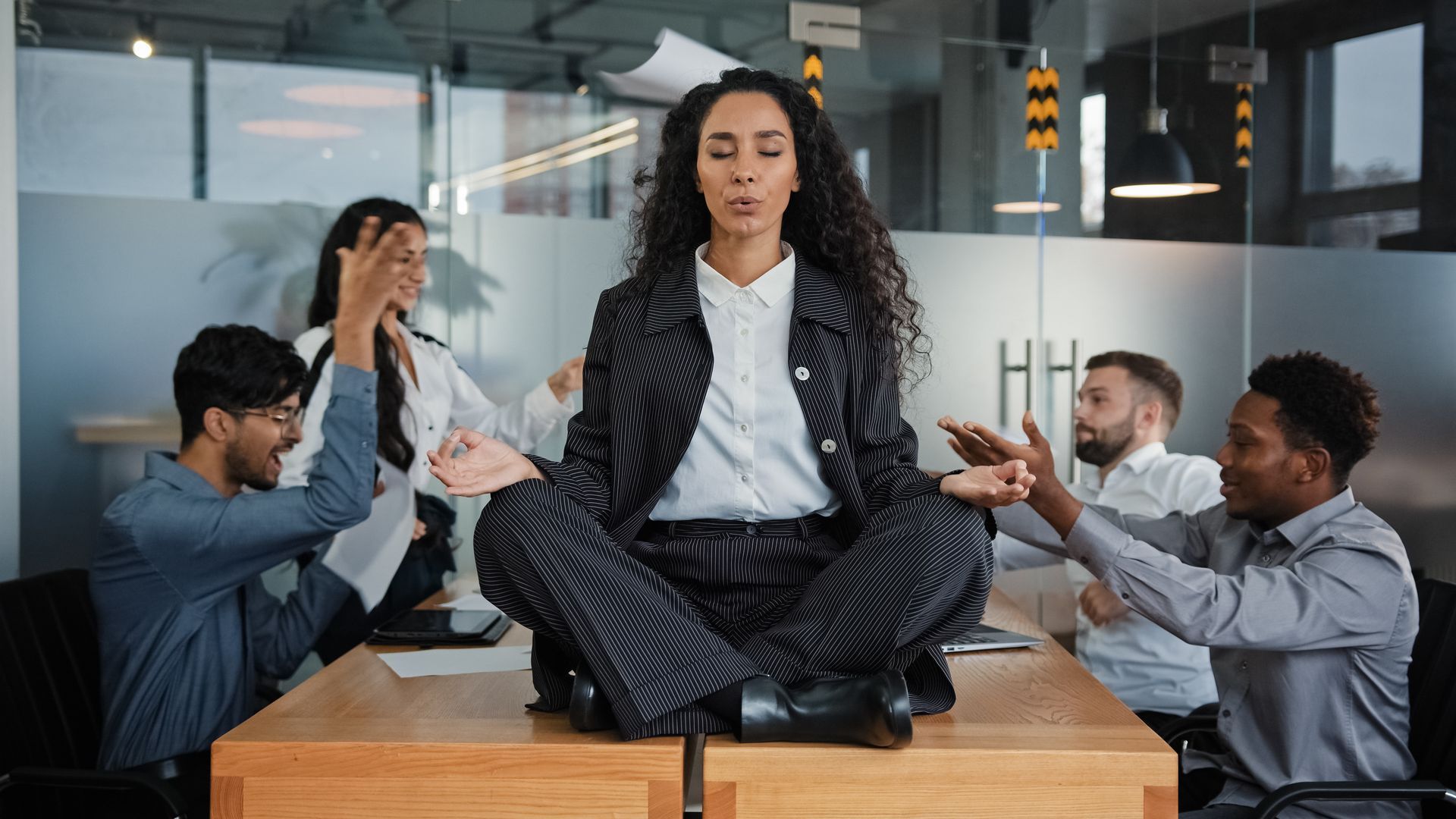 Imagem de uma mulher meditando enquanto seus colegas de trabalho discutem
