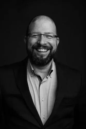 Um homem com barba e óculos sorri em uma foto em preto e branco.