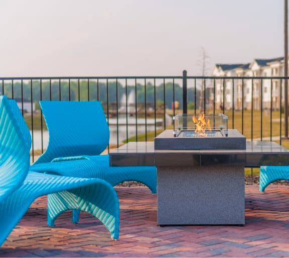Two turquoise chairs face a lit fire pit on a brick patio overlooking water and buildings.