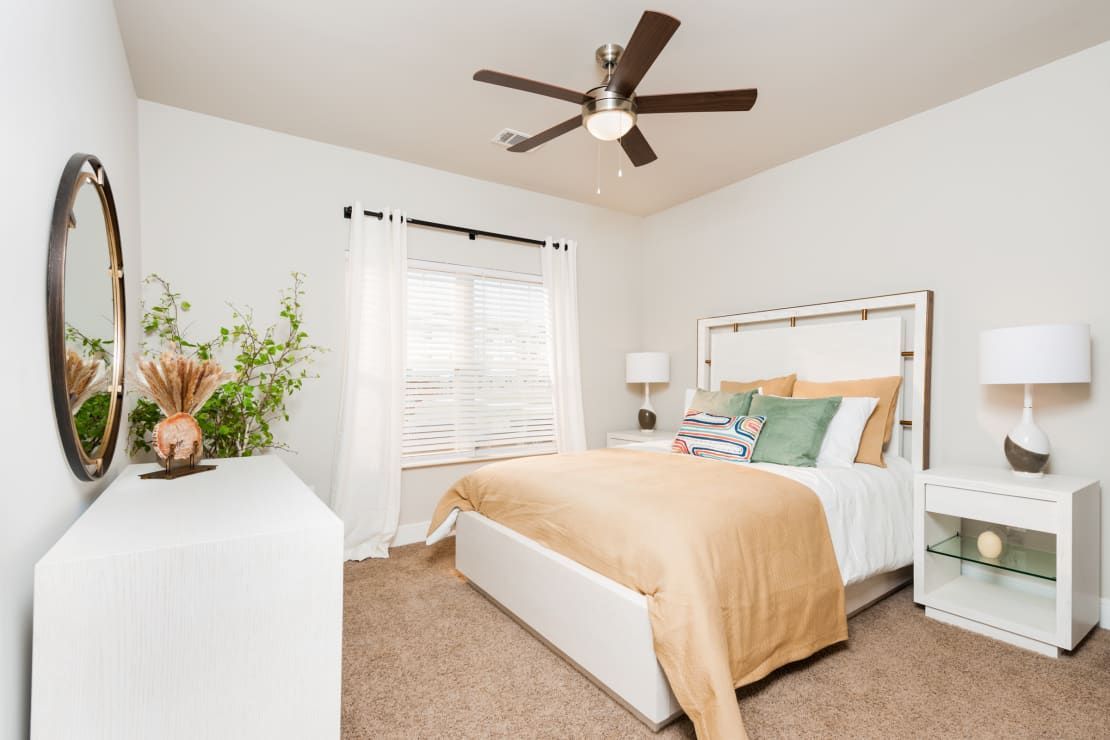 Bedroom with bed, white dresser, mirror, and white nightstand. Brown carpet. Light colors.
