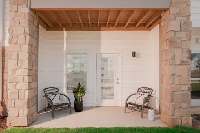 Covered patio with stone pillars, two chairs, door, and potted plant.