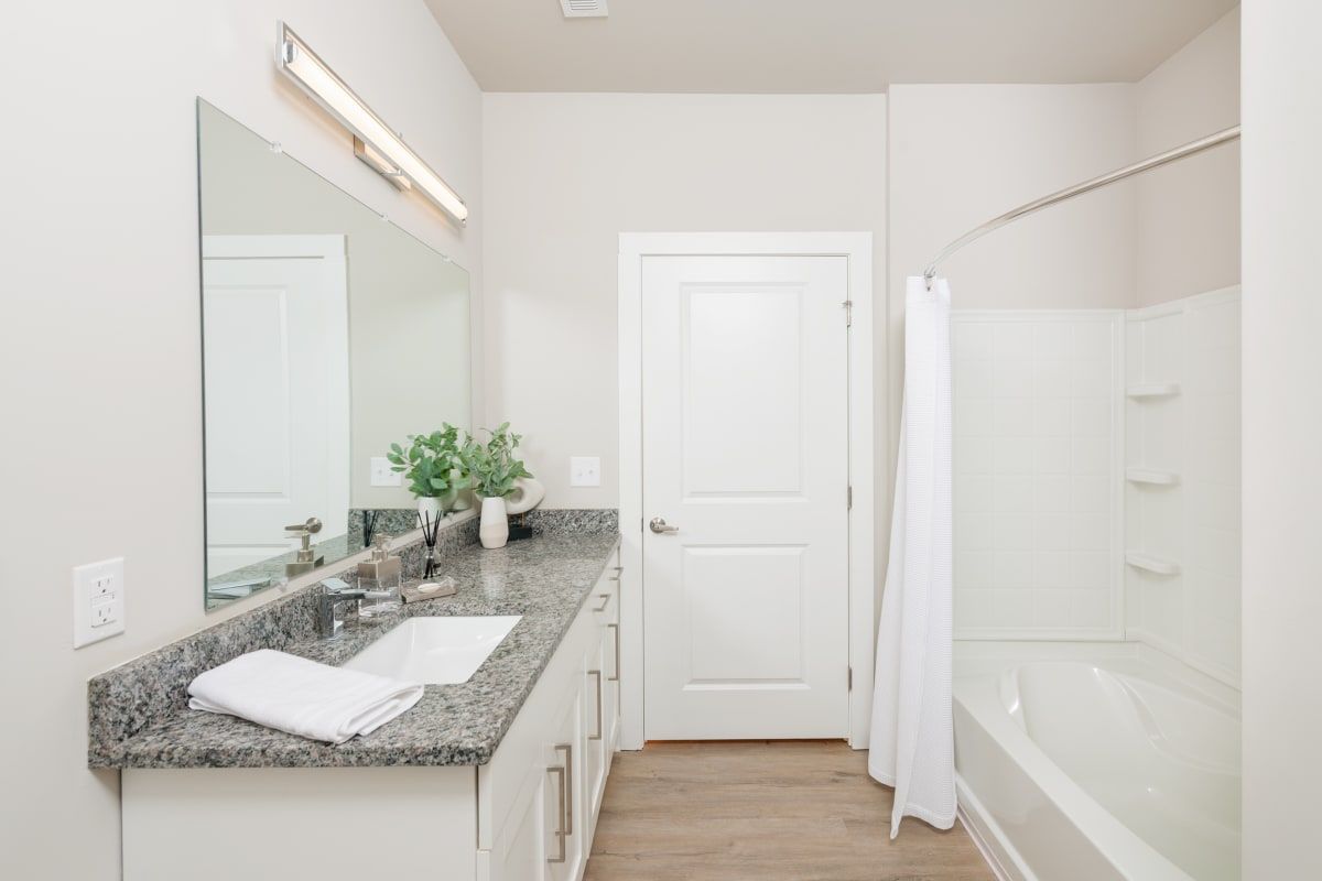 Bathroom with granite countertop, white cabinets, tub with shower curtain, and a large mirror.