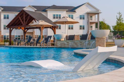 Swimming pool with white chaise lounge in water, cabana with seating, and apartment building in background.