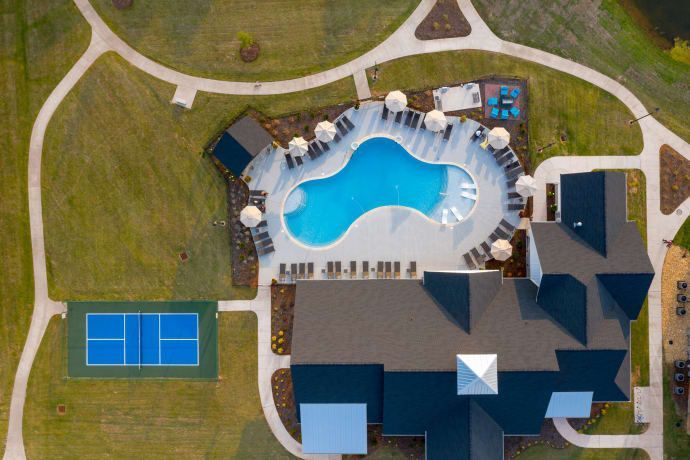 Aerial view of a community pool with lounge chairs, a building, a tennis court, and walking paths.