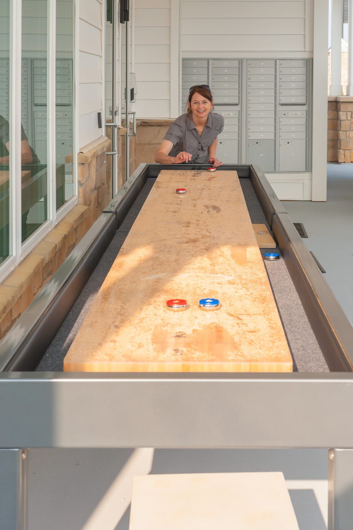 Woman playing shuffleboard outdoors, smiling. Brown wooden table, blue/red pucks.
