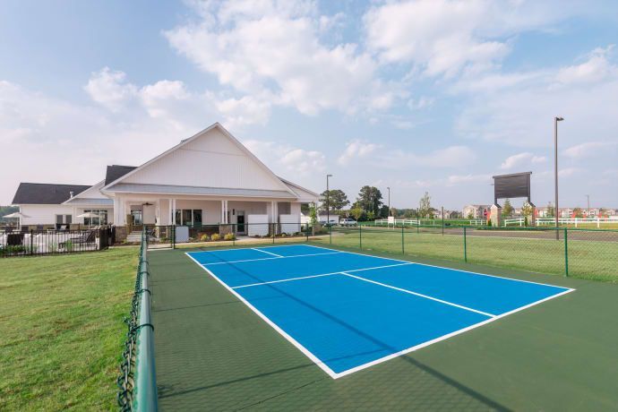 Blue pickleball court with white lines, green surrounding field, and a white building in the background.