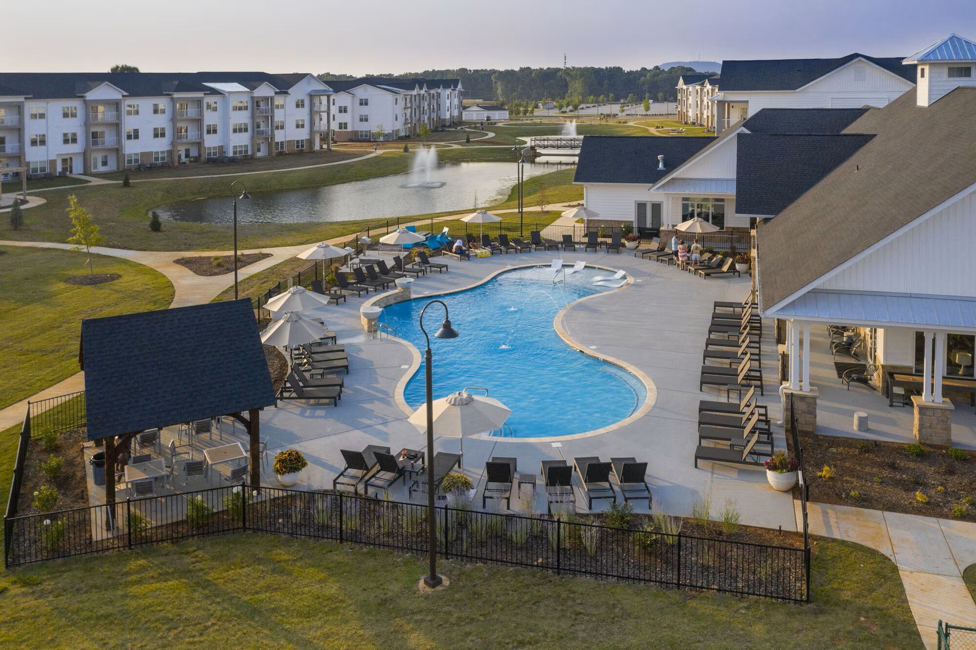 Swimming pool with lounge chairs, surrounded by buildings and a pond on a sunny day.