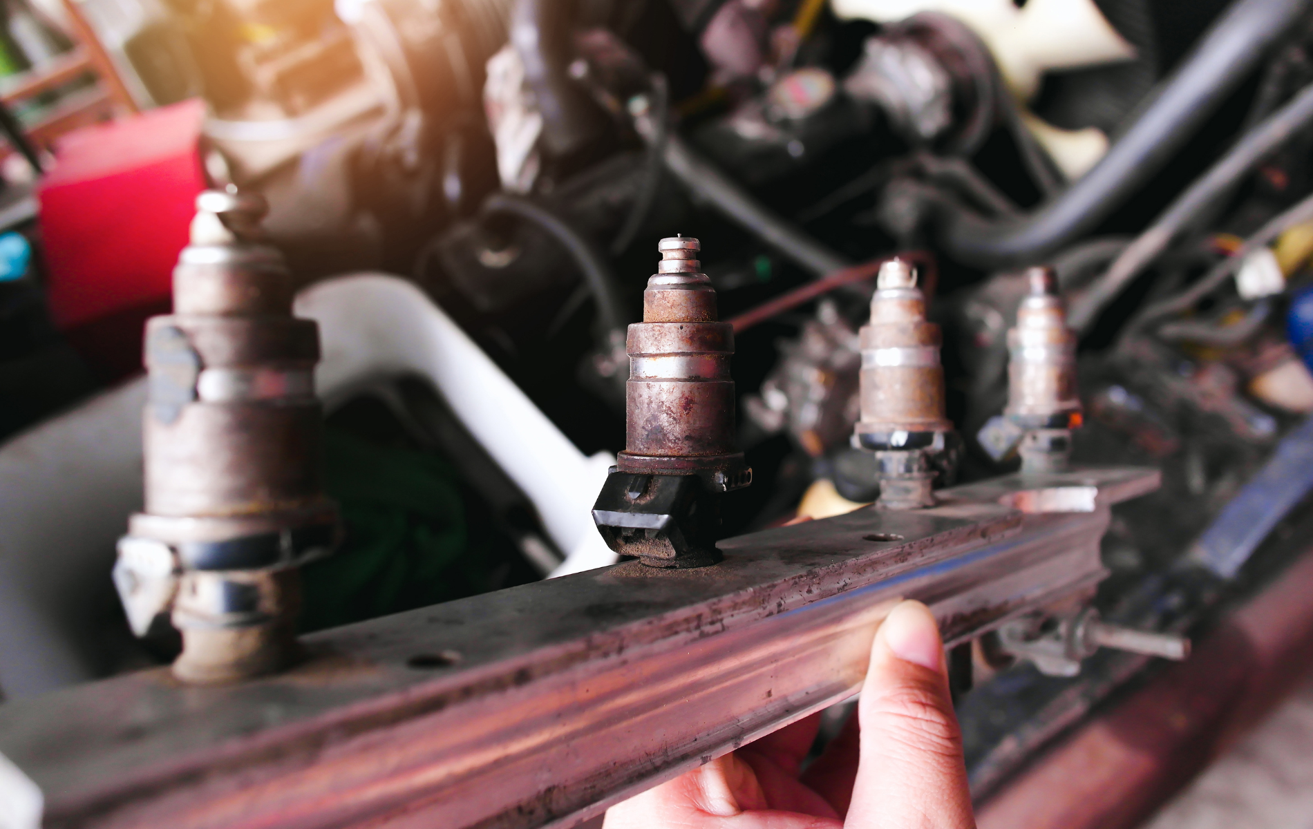 Close-up of fuel injectors on a metal rail being held with a hand, engine bay in the blurred background.
