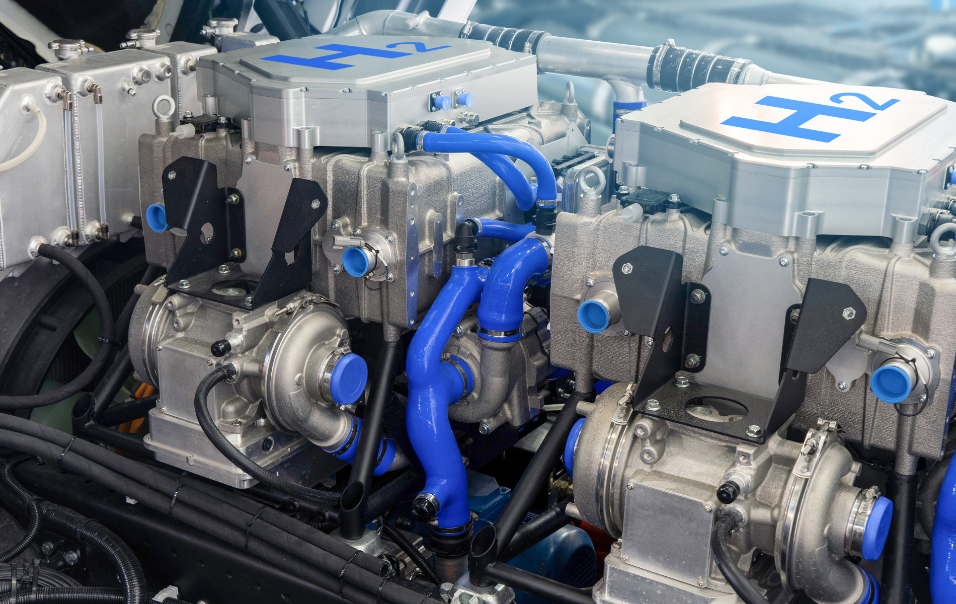 Close-up of two hydrogen fuel cell stacks with blue pipes and H2 symbols, in an engine bay.