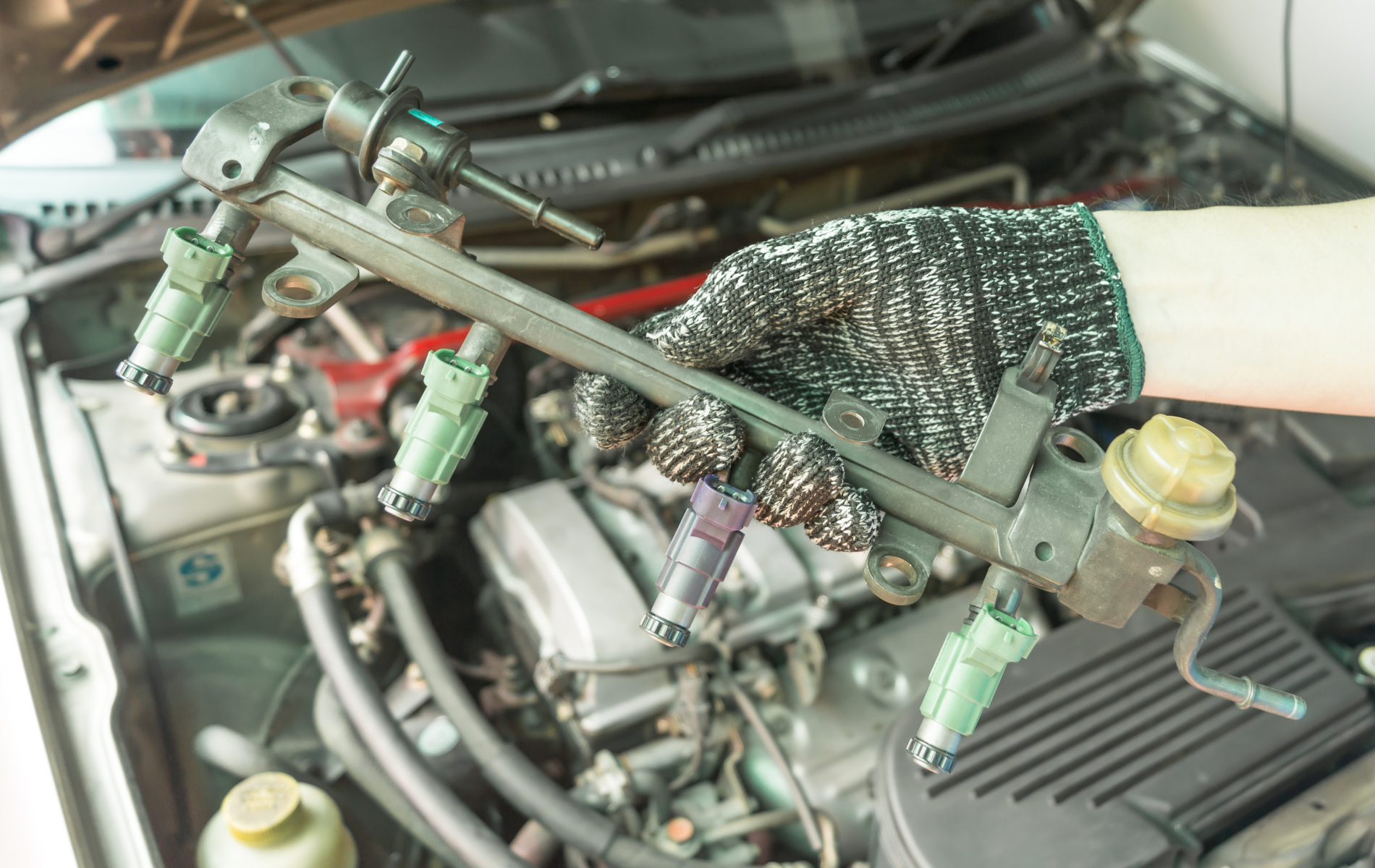 Gloved hand holding a fuel rail with four injectors over an open car engine.