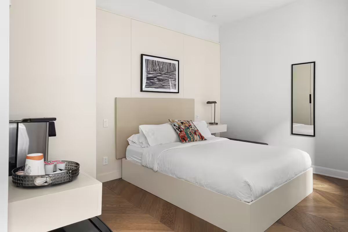 A modern hotel bedroom with a queen bed, beige headboard, framed wall art, a mirror, and a coffee station on a shelf.