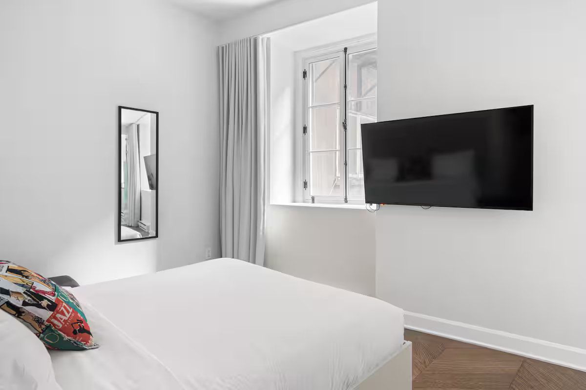 A clean, bright bedroom with white walls, a bed, a wall-mounted TV, a floor-length mirror, and window curtains.