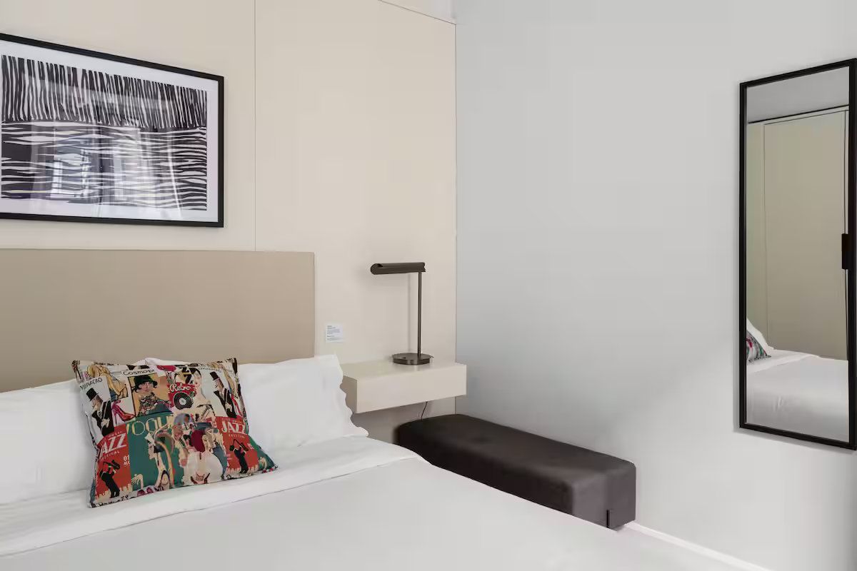 A bedroom featuring a bed with a patterned pillow, a wall-mounted bedside shelf with a lamp, and a full-length wall mirror.