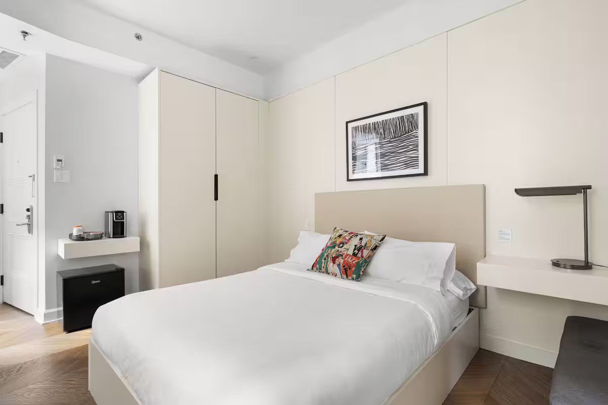 A contemporary bedroom featuring a neutral color palette, a bed with white linens, a tall wardrobe, and a desk area.
