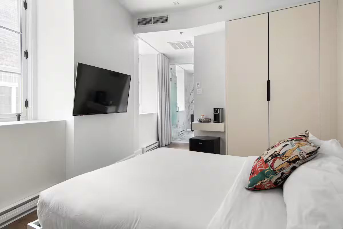 A bright, modern hotel room with a white bed, a wall-mounted television, and a cream-colored wardrobe.