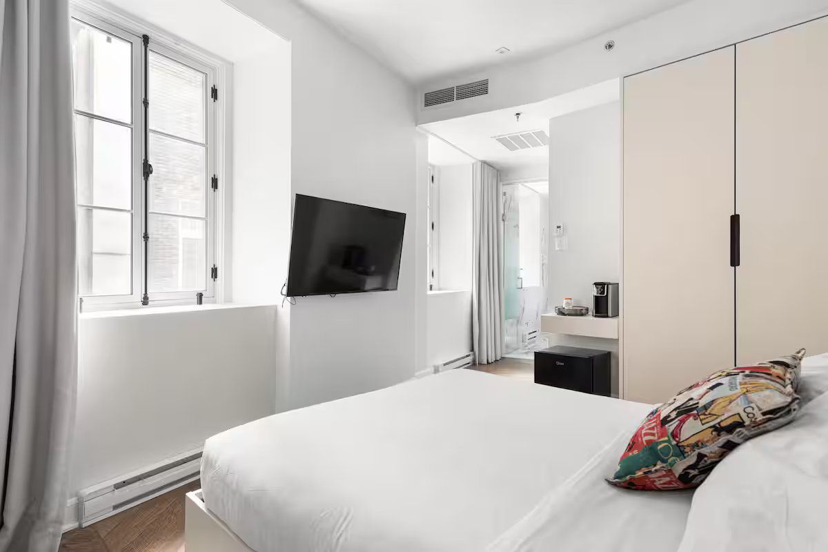 A hotel room with a bed, a wall-mounted TV, a window with grey curtains, and a light-colored wardrobe.