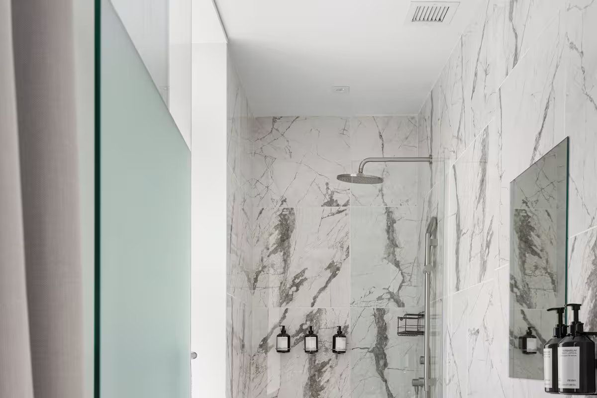A modern shower with white marble walls, a rainfall showerhead, and soap dispensers mounted on the wall.