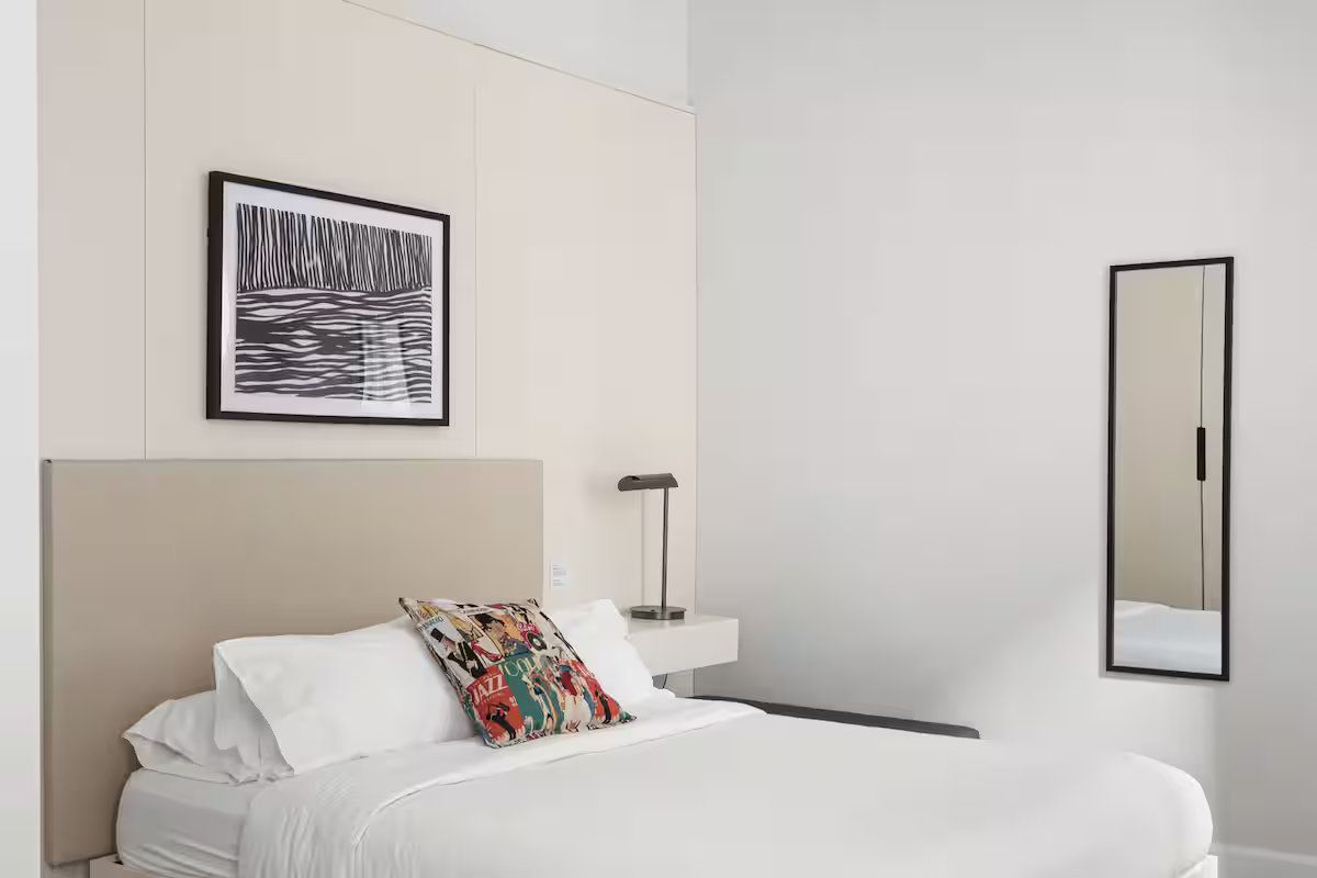 A modern hotel room with a neatly made bed, beige headboard, framed abstract art, bedside lamp, and a full-length mirror.