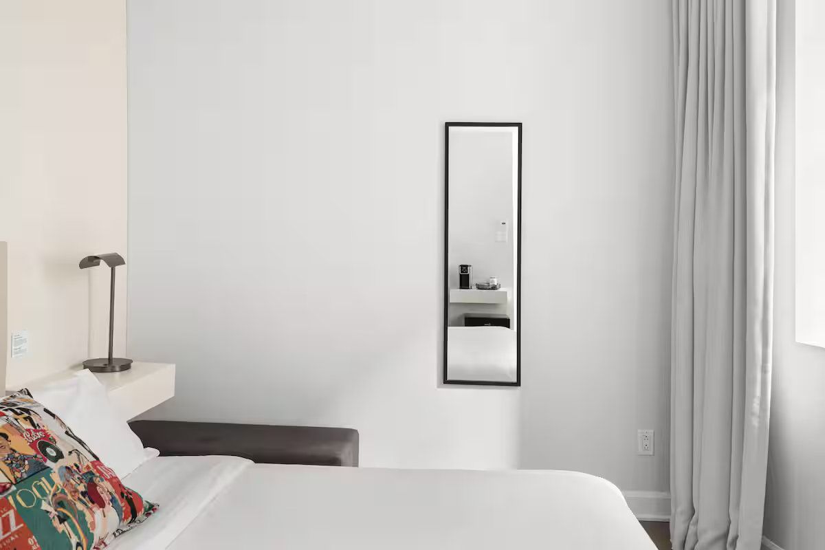 A bedroom featuring a bed with a colorful pillow, a wall-mounted rectangular mirror, and light gray curtains.