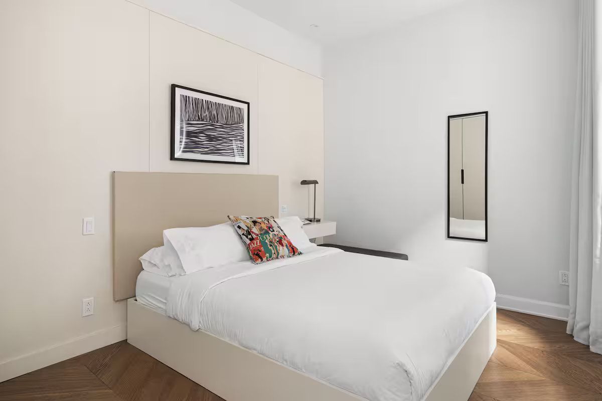 A minimalist bedroom with a cream upholstered bed, white bedding, a decorative pillow, wall art, and a vertical mirror.
