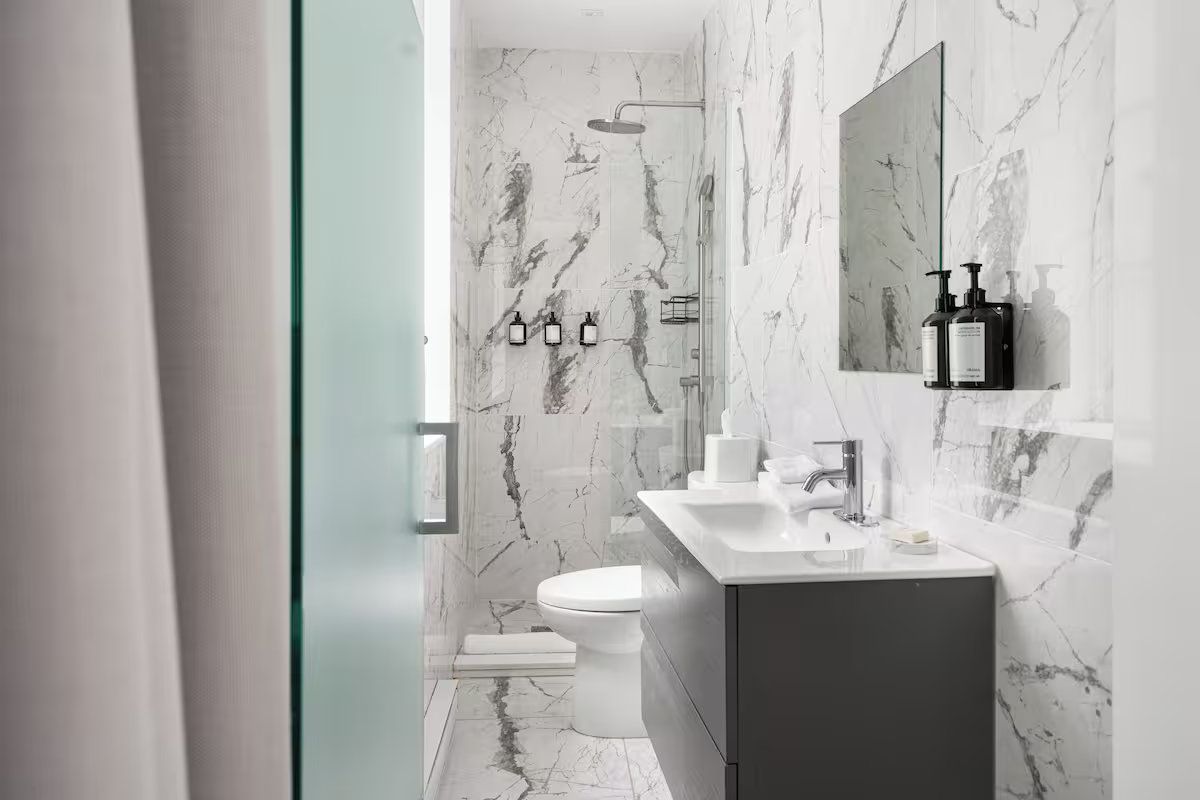 A modern bathroom with white marble walls, a gray vanity, and a toilet, seen through a partially open glass door.