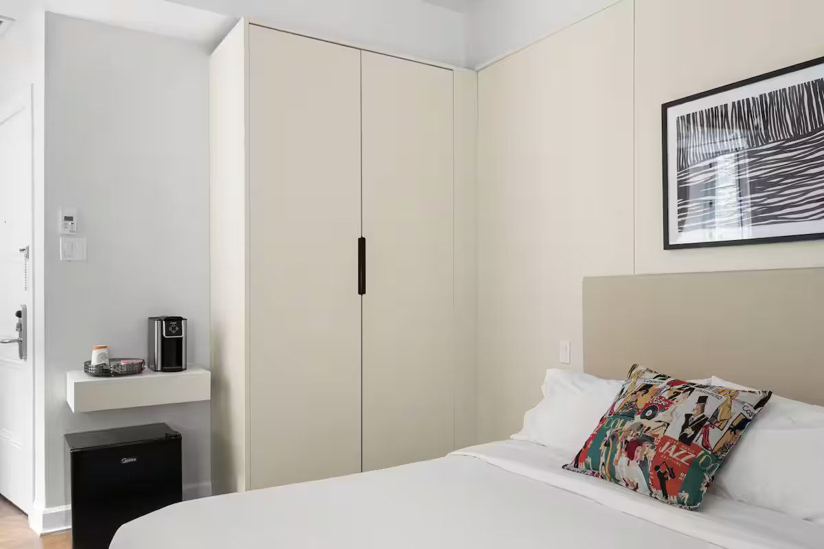 A bright, minimalist hotel room featuring a white wardrobe, a bed with a colorful accent pillow, and a coffee station.