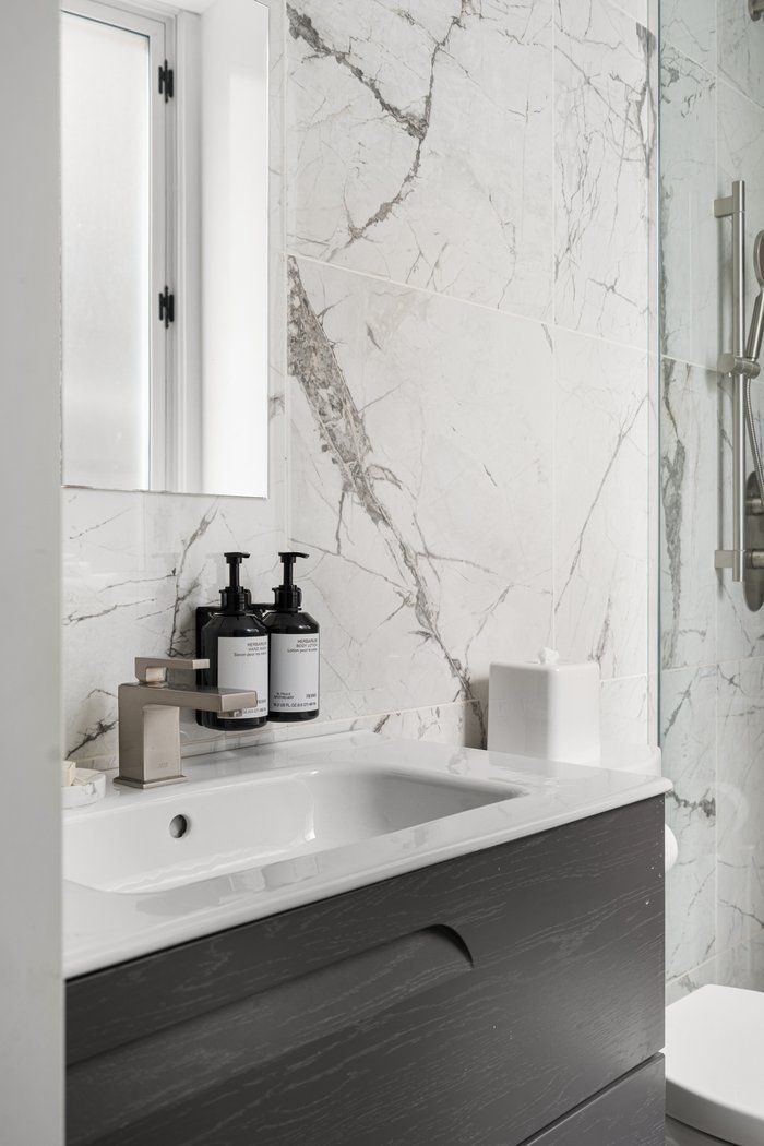 Bathroom with white marble wall, gray cabinet, white sink, black soap dispensers, and a mirror.