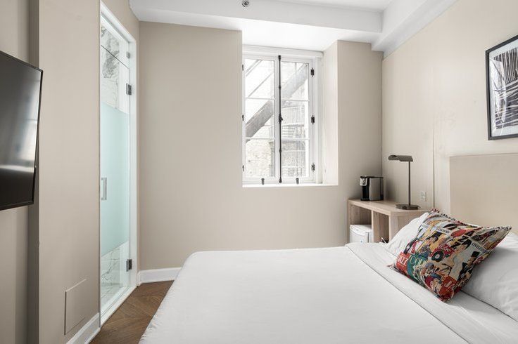 Bedroom with a bed, small desk, window, and glass shower door. Light beige walls and a patterned pillow.